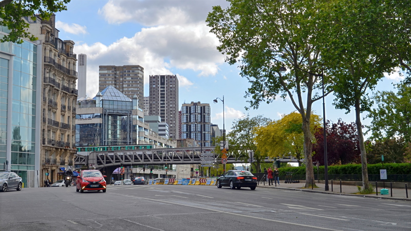 10 - An urban street scene in Paris, France, featuring a raised metro or train viaduct crossing the road amid traditional and modern buildings